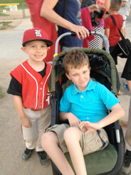 There are two brothers, one brother is wearing a red baseball uniform. The other brother is wearing a blue shirt sitting in a stroller