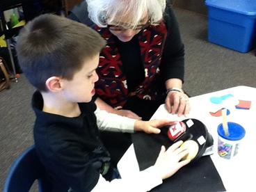 A boy and adult woman sitting at his table. He has both hands on a communication board.