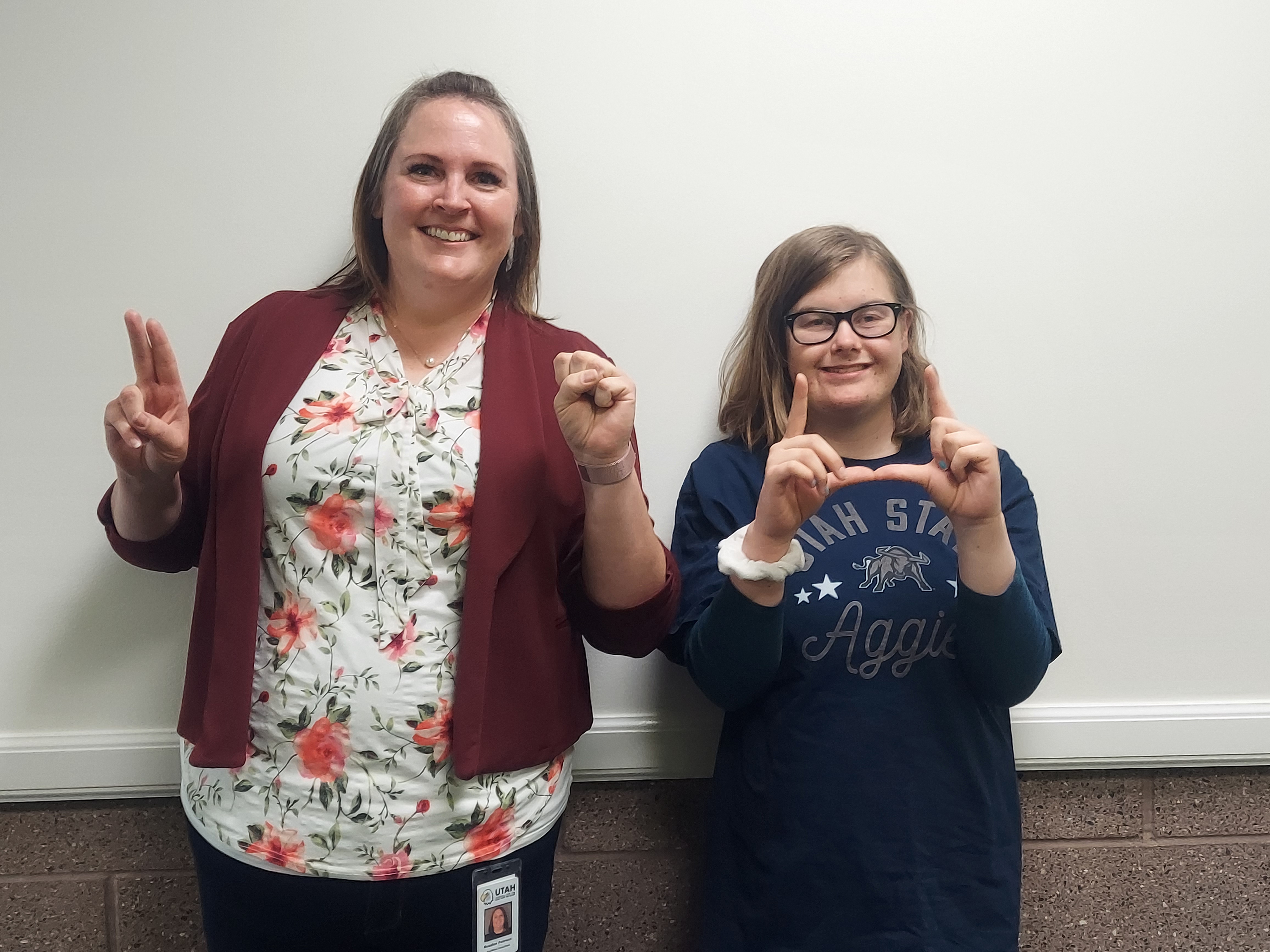 A young adult female who is deafblind stands next to an older adult female. Together they sign the letters USU for Utah State University.