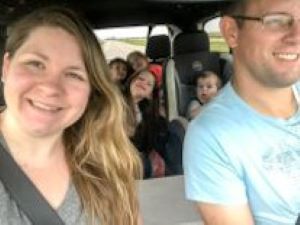 Family in car driving down a rural road. Mom and dad are in the front of the car, kids in the back. All smiling.