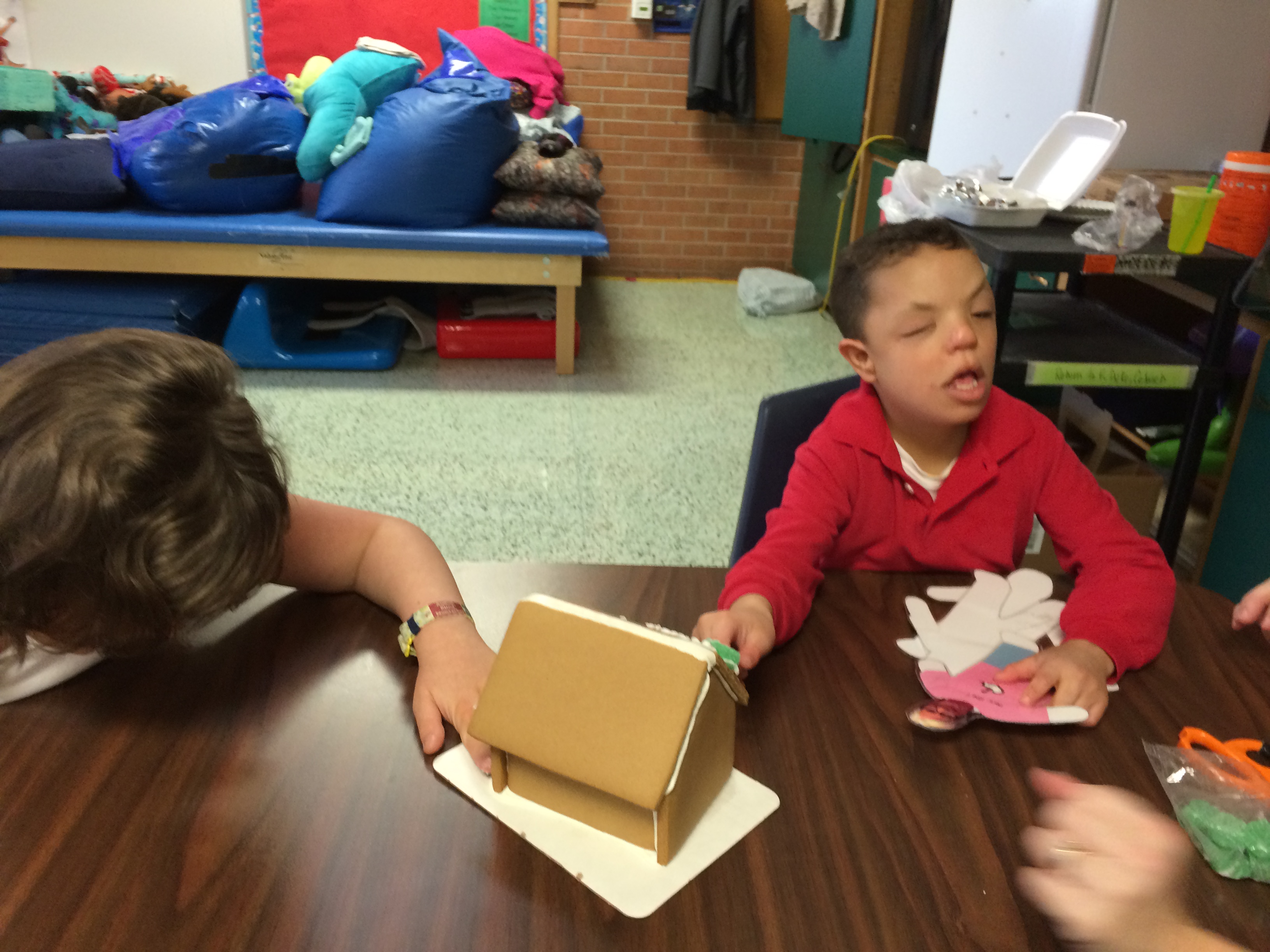 Two children sit at a table and build a gingerbread house.