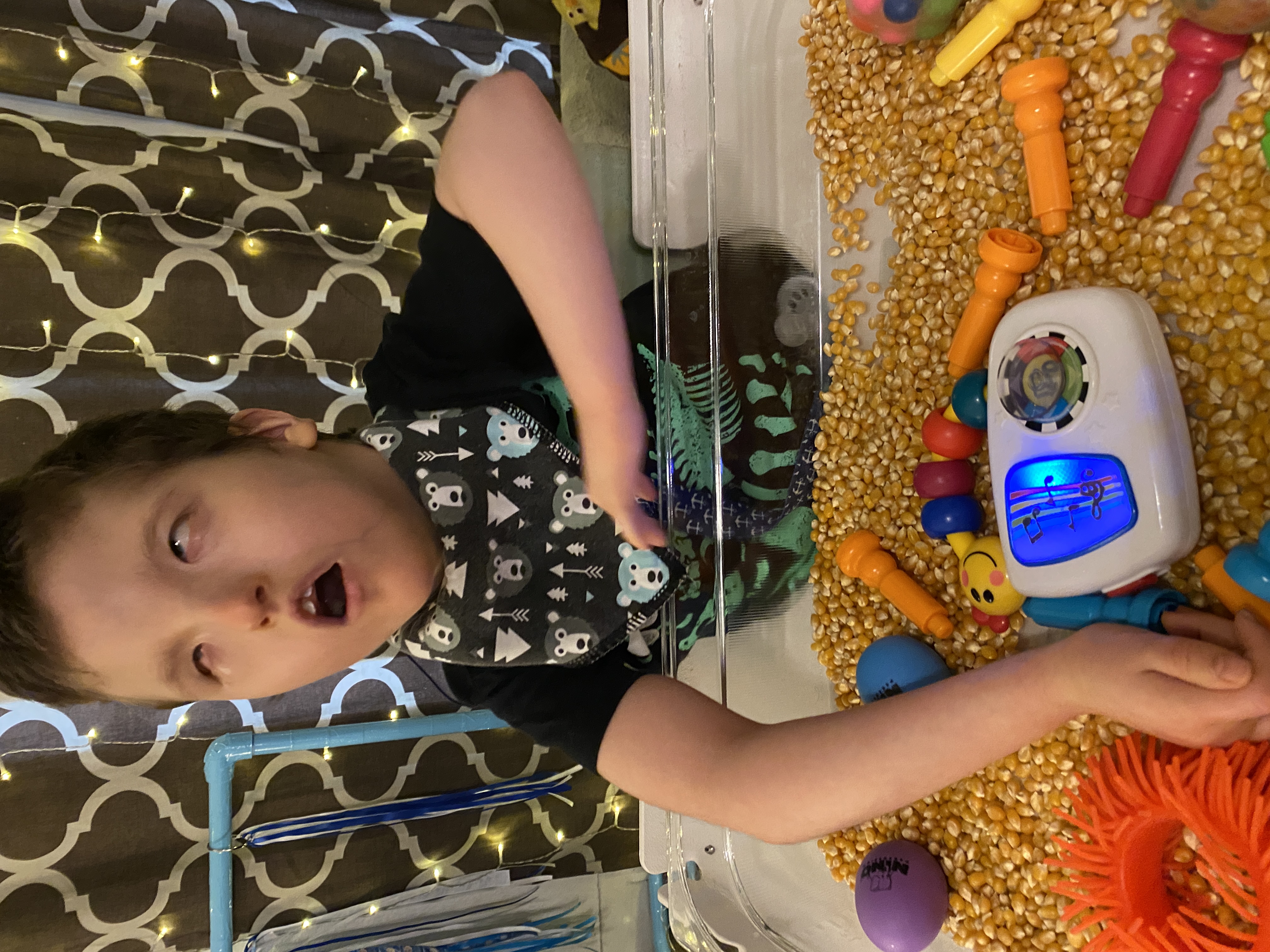 A boy with a visual impairment, about 8 years old, handling colorful toys in a clear box. The box also contains popcorn kernels.