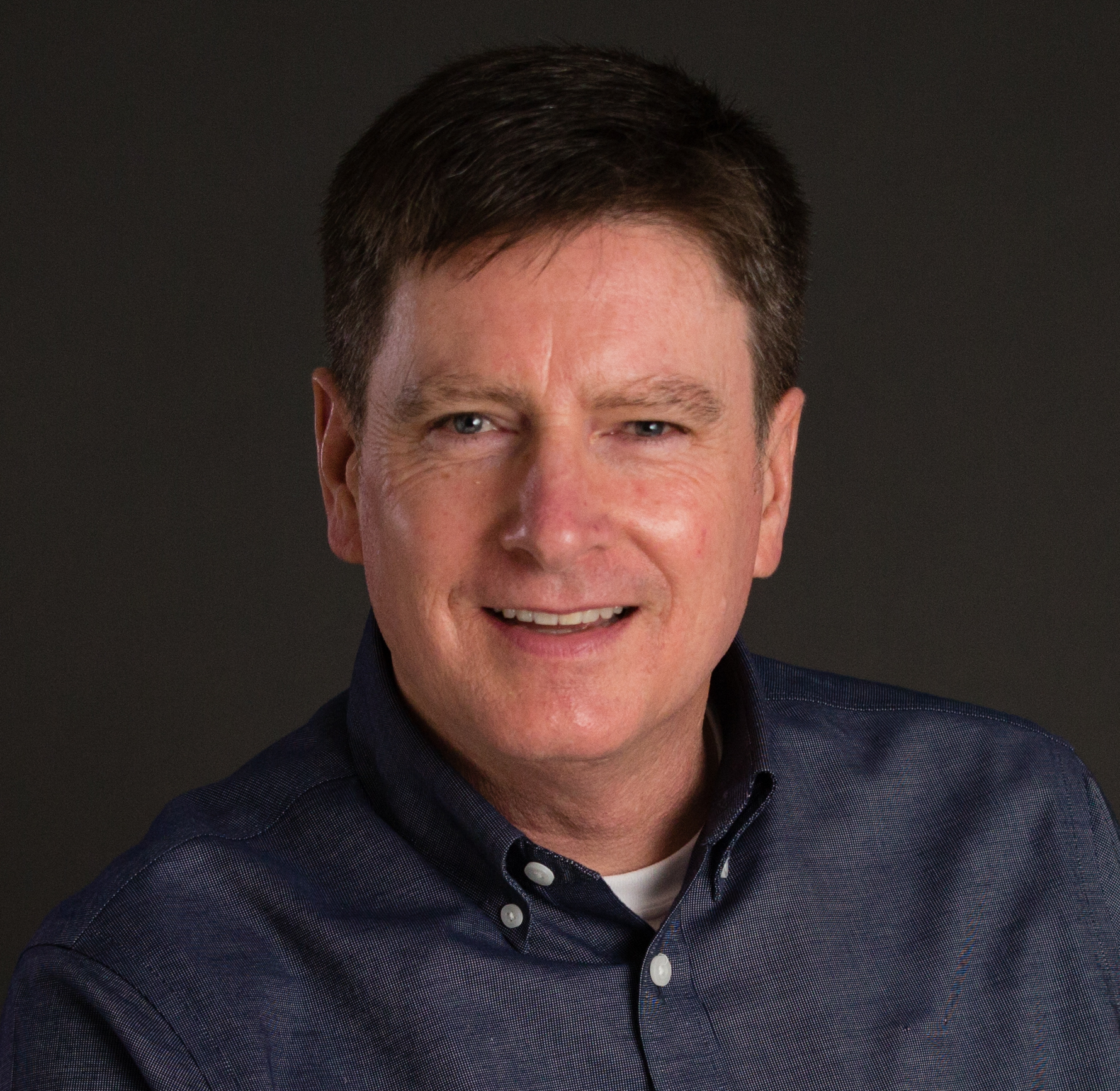 Maurice smiling with a blue button up shirt and black background.