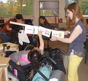 A girl who is deaf-blind is engaged in a lesson on how much things costs. She is in a wheelchair and is pointing to communication cards on which there are pictures of items and their prices.