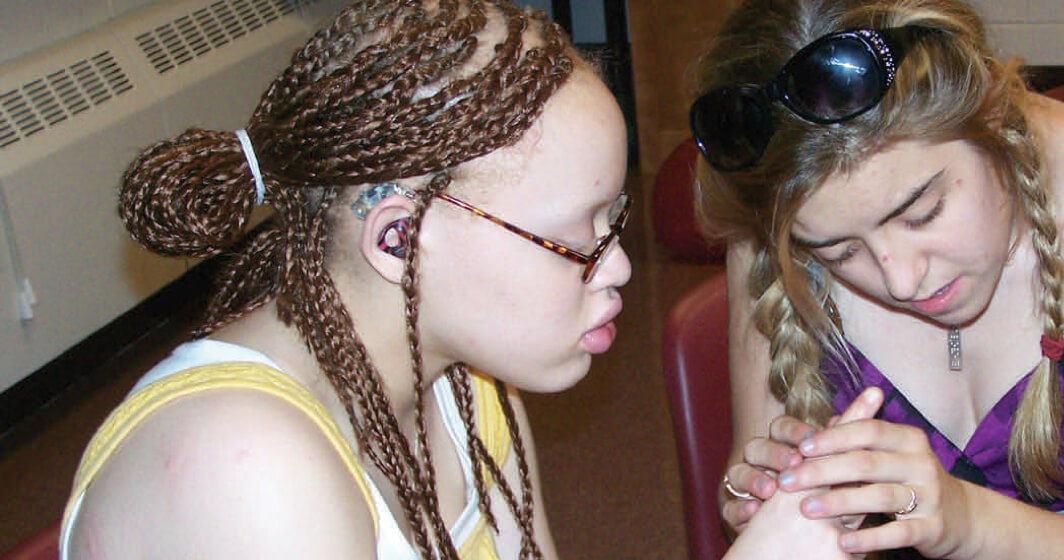 Two young women sit facing each other. They are conversing using tactile sign language.